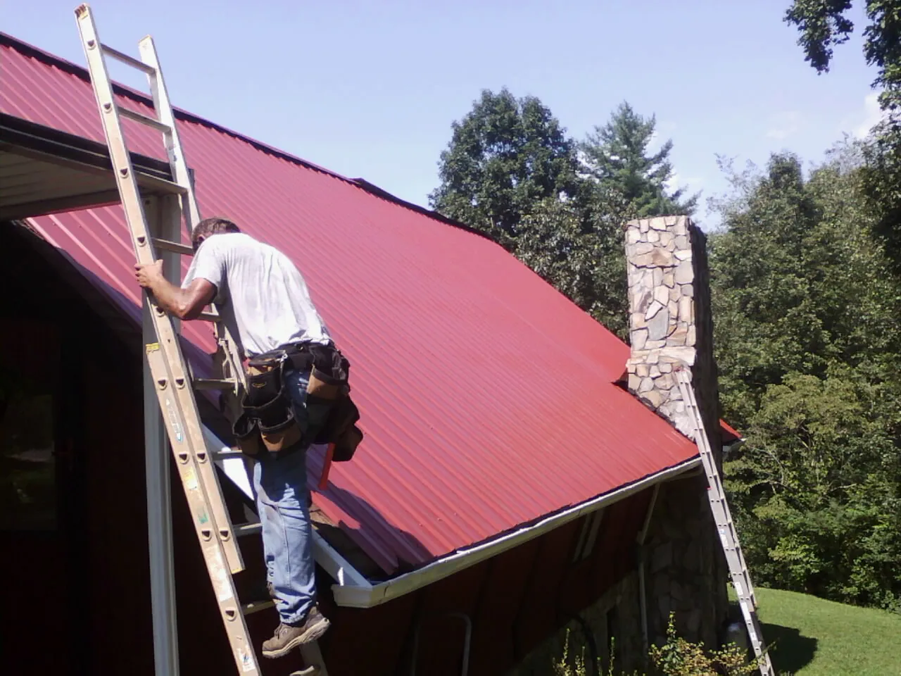 Roofer on a ladder beside a red metal roof and stone chimney — Soffit & Fascia Repair by Roofing Brothers in Bingham Canyon